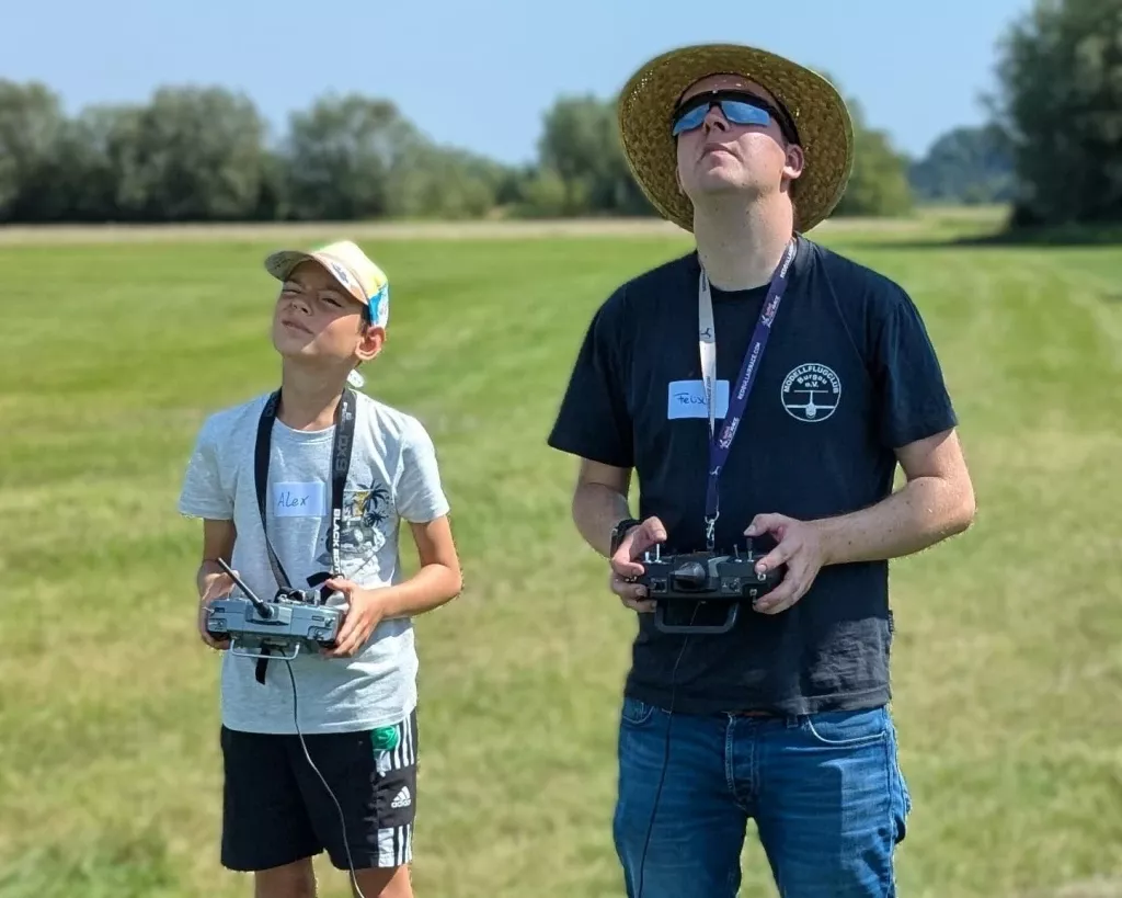 Zwei Personen mit Fernsteuerungen auf Wiese: Jugendlicher in grauem T-Shirt und erwachsener Trainer mit Strohhut und Sonnenbrille