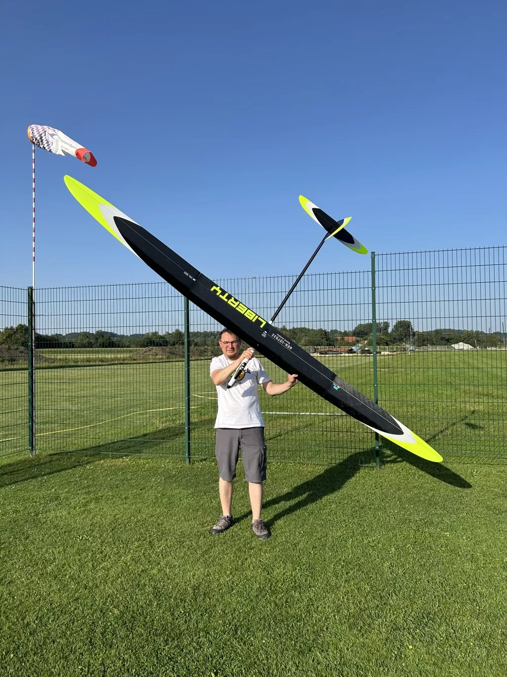 Wei&szlig;bekleidete Person h&auml;lt ein schwarzes und gelbes Segelflugmodell auf gr&uuml;nem Rasenplatz mit Windfahne