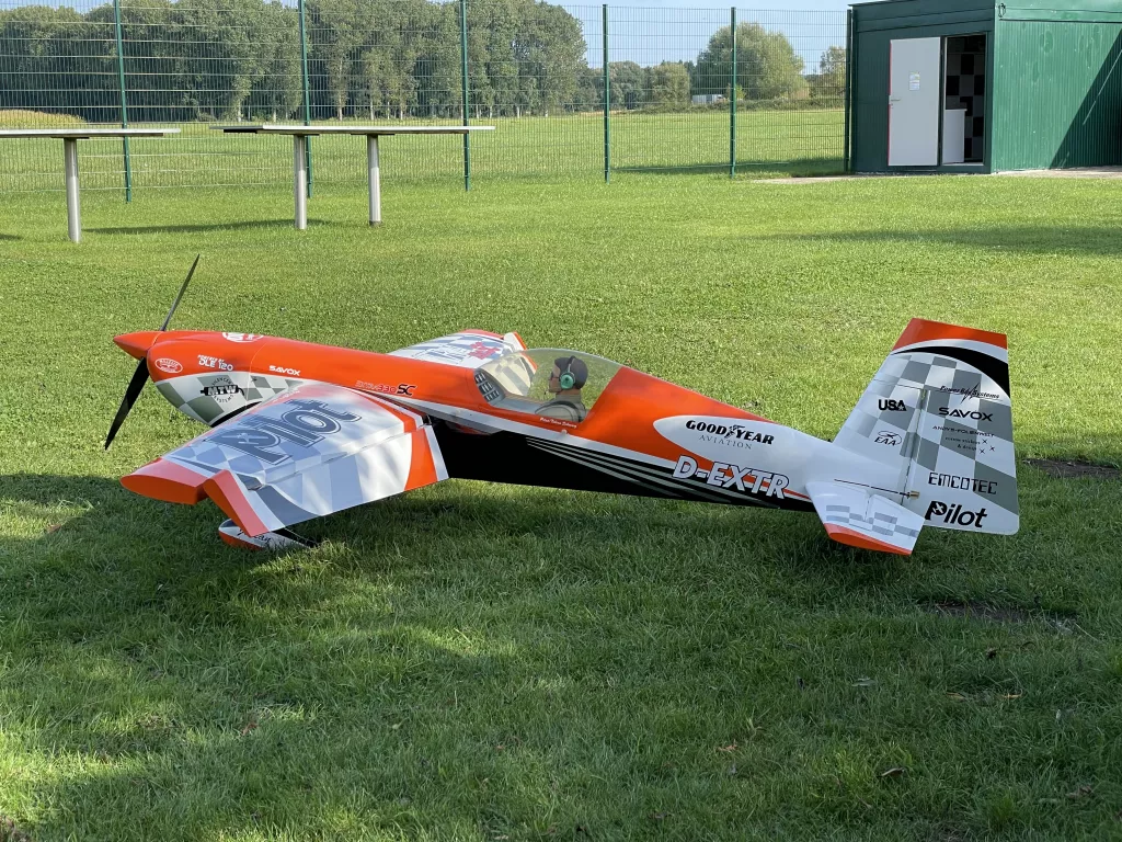 Orangewei&szlig;es Kunstflug-Modellflugzeug mit Sponsorenlogos auf gr&uuml;ner Rasenfl&auml;che vor gr&uuml;nem Container