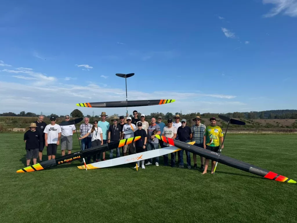 Gruppe von etwa 20 Personen mit drei gro&szlig;en Segelflugmodellen auf einer Wiese, blauer Himmel im Hintergrund