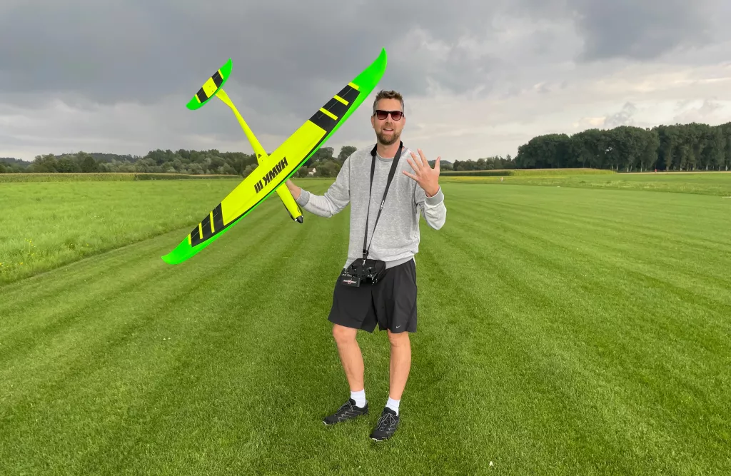 Vereinsmitglied in grauer Jacke h&auml;lt gro&szlig;es neongr&uuml;n-schwarzes Segelflugmodell auf einer gem&auml;hten Wiese mit B&auml;umen im Hintergrund