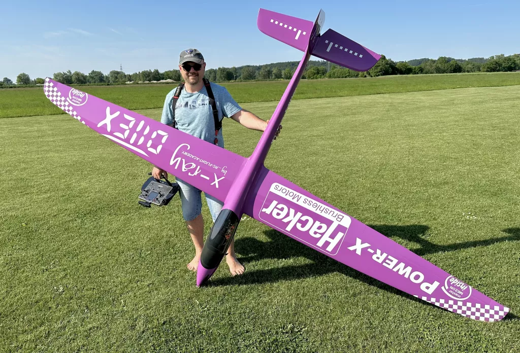 Lila Segelflugmodell X-300 auf Rasenfl&auml;che, daneben ein Modellflug-Enthusiast mit Basecap und Sonnenbrille, der eine Fernsteuerung h&auml;lt