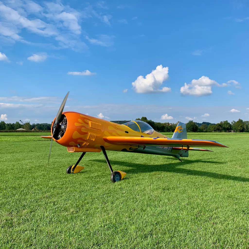 Knallorange-gelbes Kunstflugmodell mit Propeller steht auf gr&uuml;ner Rasenfl&auml;che vor blauem Sommerhimmel
