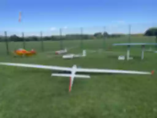 Mehrere Modellflugzeuge auf grüner Rasenfläche, darunter ein gelbes Motorflugzeug und weiße Segelflugzeuge, umgeben von einem Maschendrahtzaun