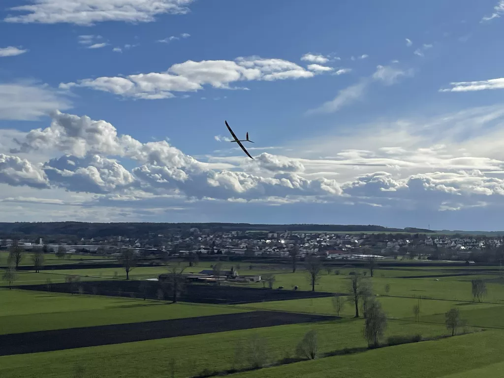Schlankes Segelflugmodell fliegt &uuml;ber weite Feldlandschaft mit gr&uuml;nen Wiesen und Dorf im Hintergrund, blauer Himmel mit Wolken