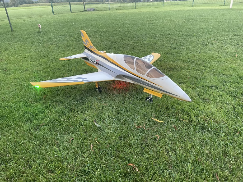 Wei&szlig;es und gelbes Modellflugzeug mit Strahlertriebwerk auf gr&uuml;ner Rasenfl&auml;che, Vereinsgel&auml;nde mit Zaun im Hintergrund