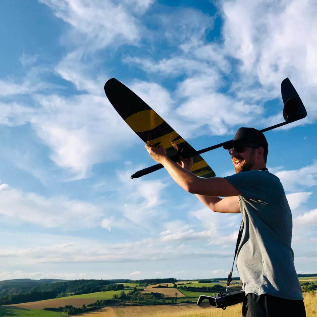 Person h&auml;lt gestreiftes Segelflugmodell in gelb-schwarzer Farbe vor blauem Himmel und gr&uuml;ner Landschaft