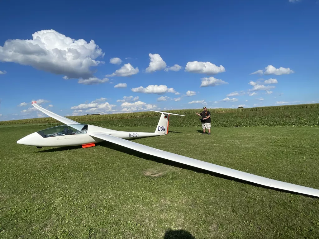 Wei&szlig;es Segelflugzeugmodell mit rot-wei&szlig;er Kennung auf gr&uuml;ner Wiese, im Hintergrund Felder und Himmel