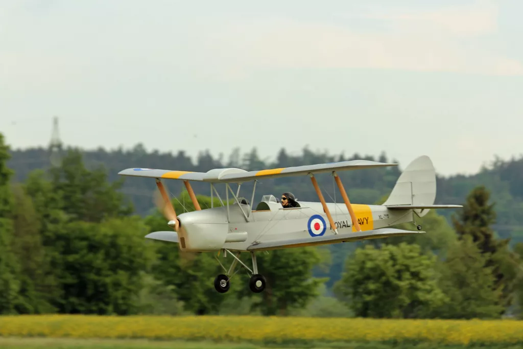 Wei&szlig;er Doppeldecker im Flug, gelbe Fl&uuml;gelkanten, Royal Navy Abzeichen, Pilot sichtbar, Nadelwald im Hintergrund