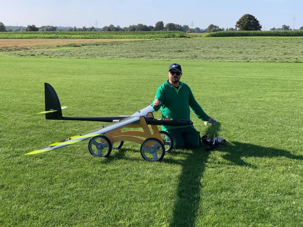 Modellflugzeug auf gr&uuml;ner Wiese, wei&szlig;-gelb-golden, mit R&auml;dern und schwarzer Fl&uuml;gelspitze, Person in gr&uuml;nem Outfit daneben
