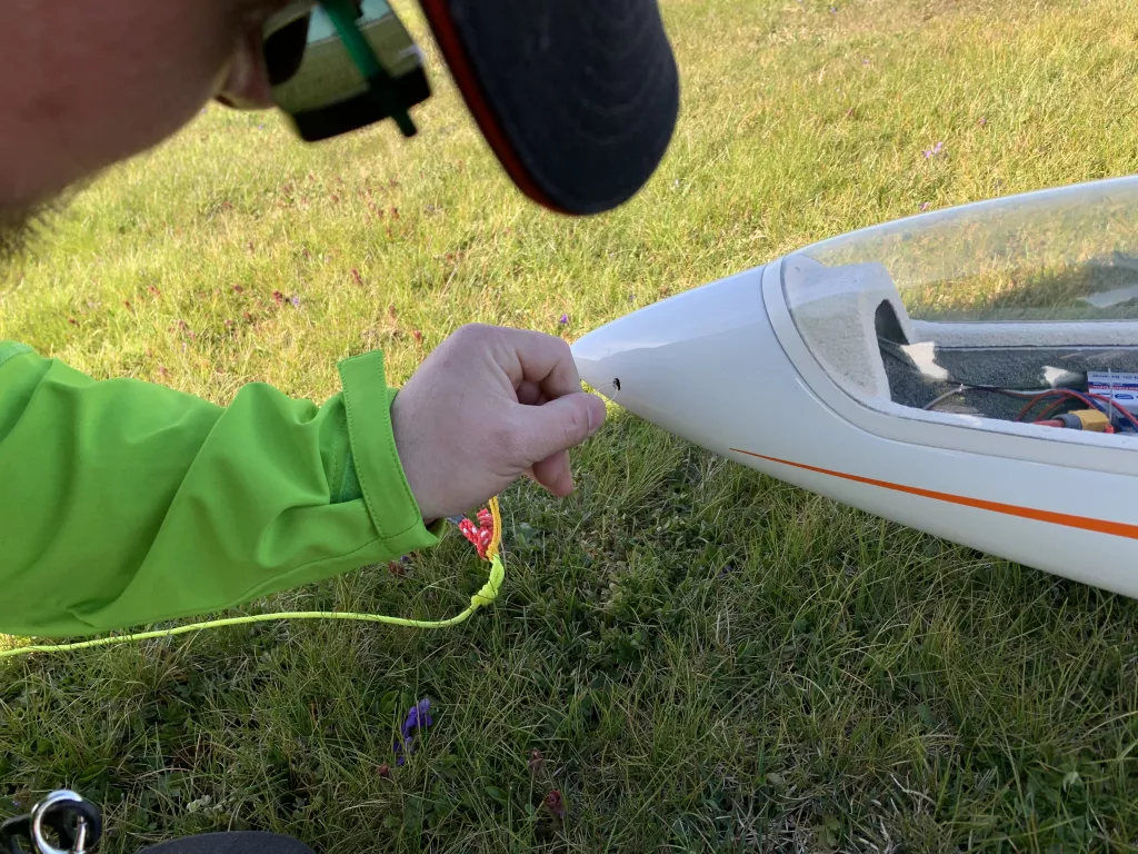 Eine Hand in gr&uuml;ner Jacke h&auml;lt ein wei&szlig;es Segelflugmodell auf einer Wiese mit gelbgr&uuml;nem Gras