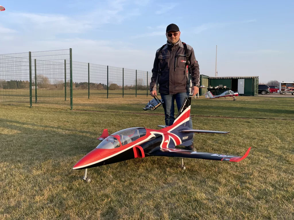 Modellflugzeug im Vereinsfarben Rot-Blau-Wei&szlig;, daneben ein Modellpilot in grauer Jacke auf Rasenfl&auml;che mit Zaun