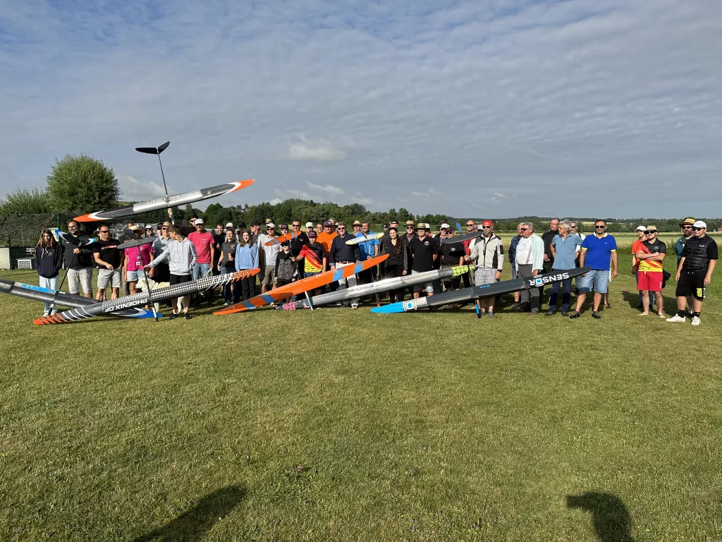 Gro&szlig;e Gruppe von Modellflugbegeisterten mit verschiedenfarbigen Segelflugmodellen auf gr&uuml;ner Wiese unter bew&ouml;lktem Himmel