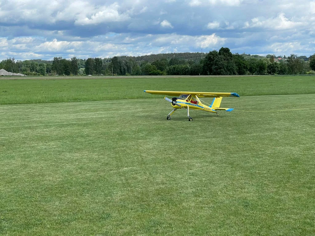 Gelb-blaues Modellflugzeug steht auf gr&uuml;ner Rasenfl&auml;che mit B&auml;umen und bew&ouml;lktem Himmel im Hintergrund