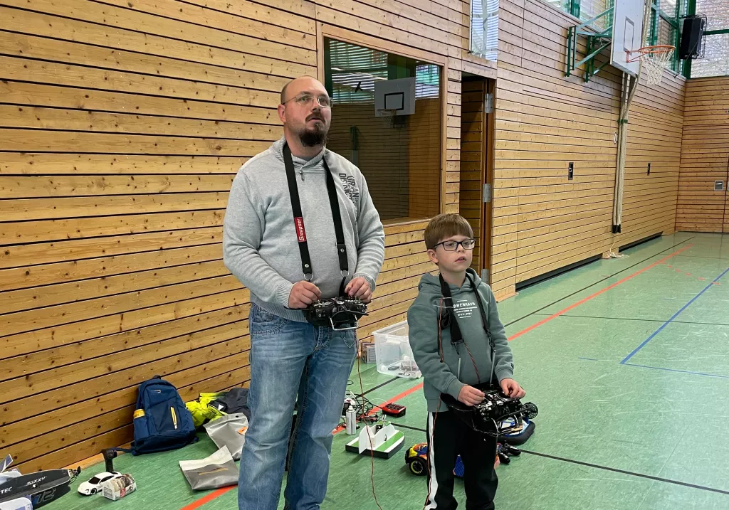 Vater und Sohn mit Fernsteuerungen in Sporthalle, umgeben von Modellfahrzeugen und Drohnen-Equipment