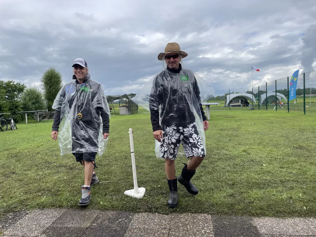 Zwei Personen in Regencapes und Gummistiefeln auf Vereinsgel&auml;nde, bew&ouml;lkter Himmel, Flaggen im Hintergrund