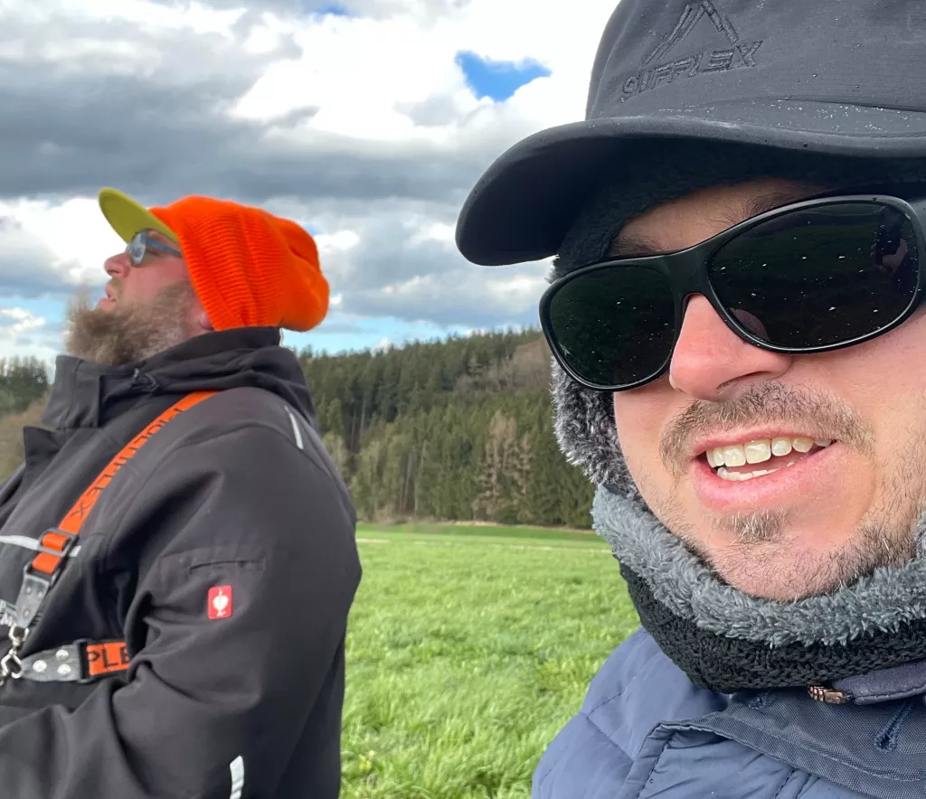 Zwei Personen in Winterkleidung auf Wiese, Wald im Hintergrund. Einer mit oranger M&uuml;tze, einer mit schwarzer Sonnenbrille und Fellkragen.