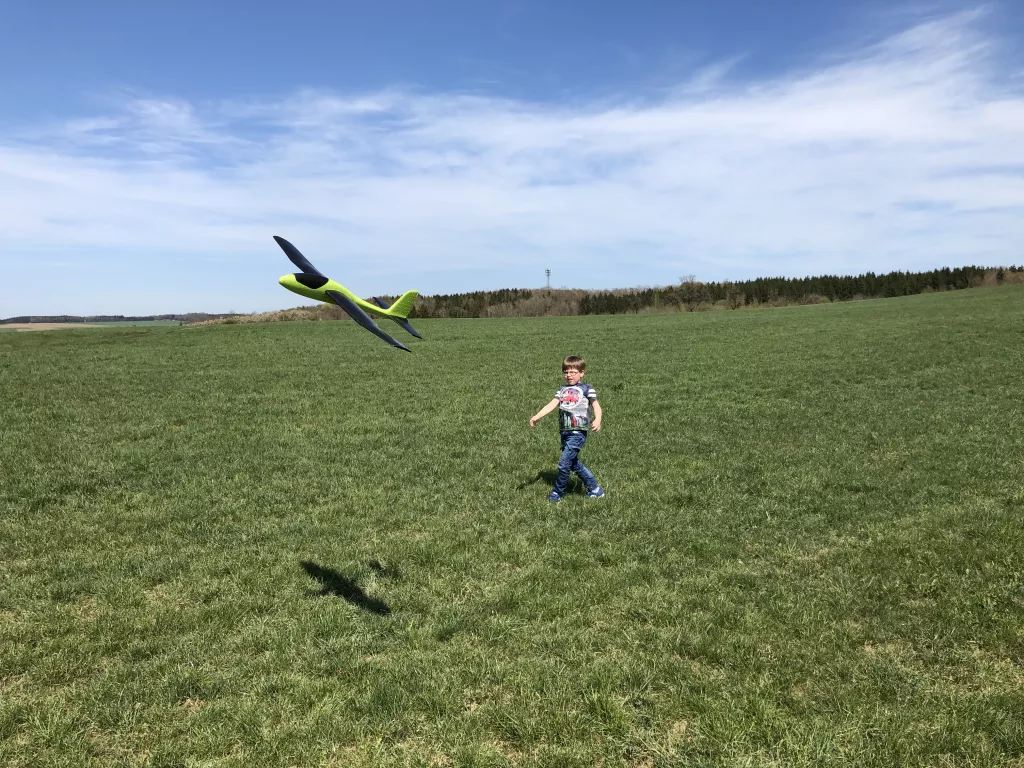Kind wirft gelb-schwarzes Segelflugmodell in blauer Landschaft, gr&uuml;ne Wiese im Vordergrund, Wald am Horizont