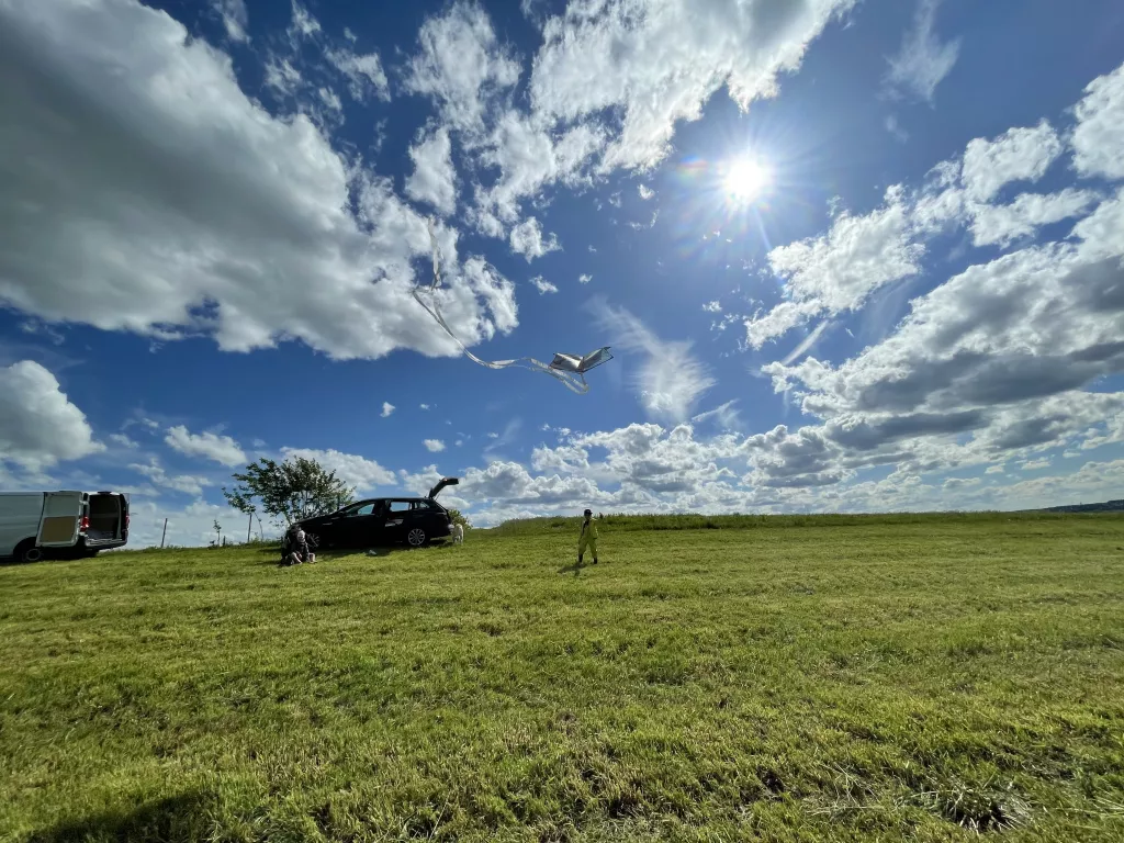 Gr&uuml;ne Wiese mit Modellflugzeug in der Luft, schwarzes Auto und Vereinsmitglied im Hintergrund, blauer Himmel mit Wolken