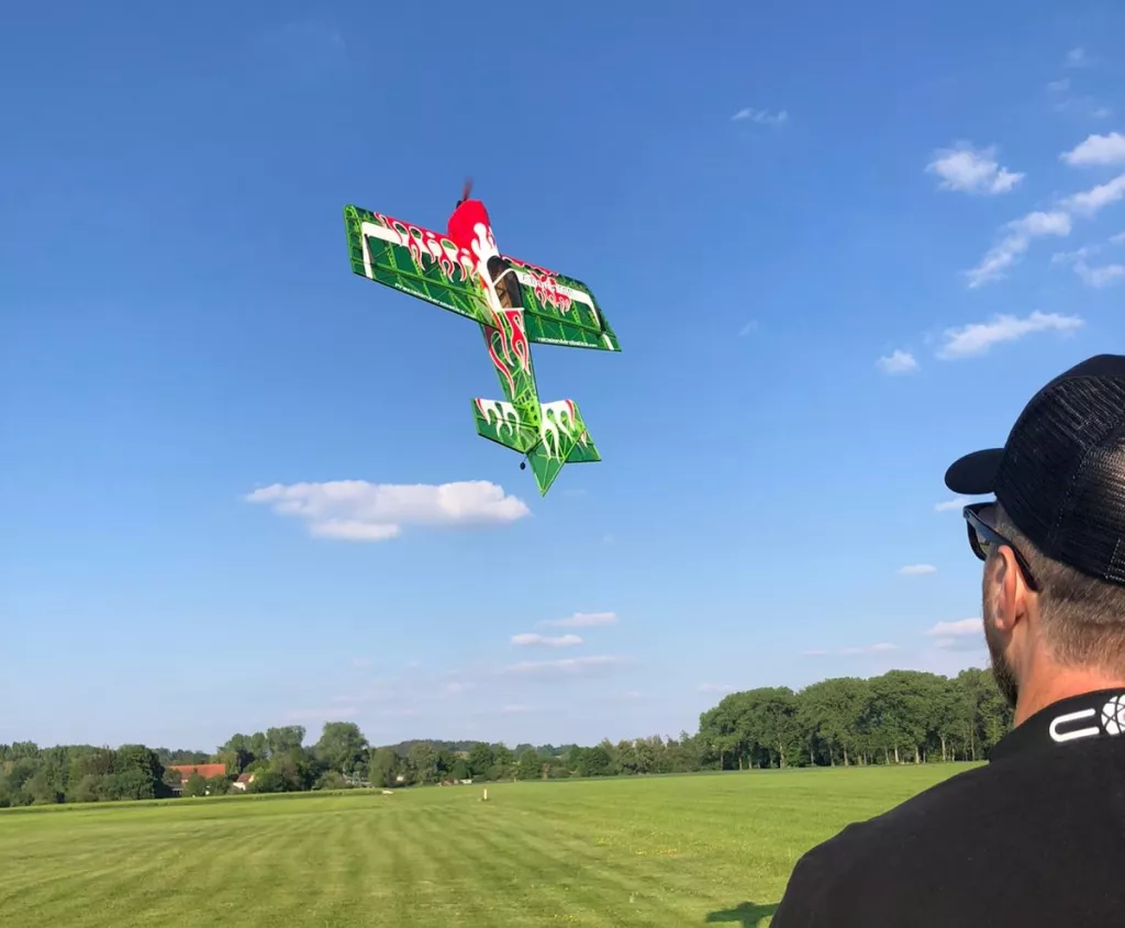 Detailreiches Modellflugzeug in gr&uuml;n-roter Lackierung w&auml;hrend eines Kunstflugs &uuml;ber eine weite gr&uuml;ne Wiese mit B&auml;umen im Hintergrund