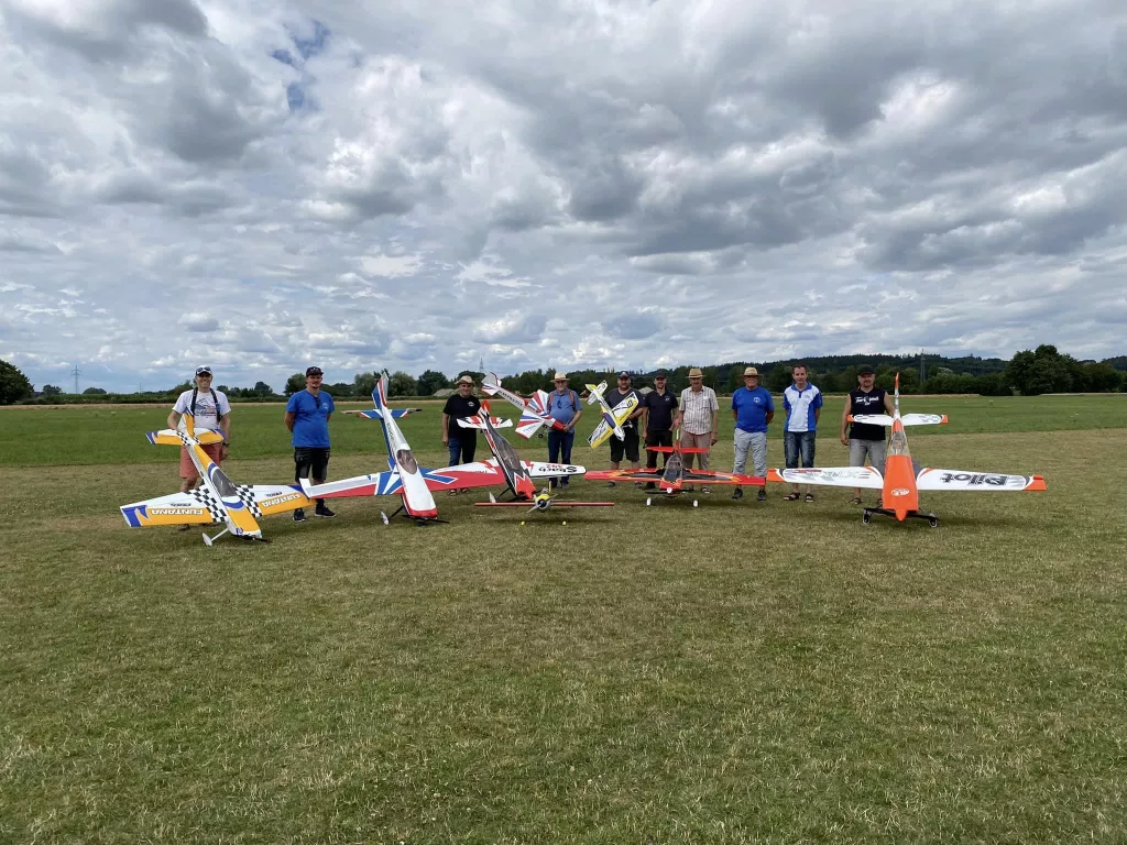 Neun Modellflugzeug-Piloten stehen mit bunten Kunstflugmodellen auf einer Wiese unter bew&ouml;lktem Himmel