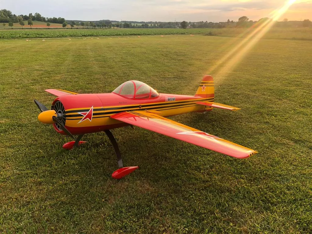 Rot-gelbes Kunstflug-Modellflugzeug auf grüner Wiese bei Sonnenaufgang, mit Propeller und Sternmotor