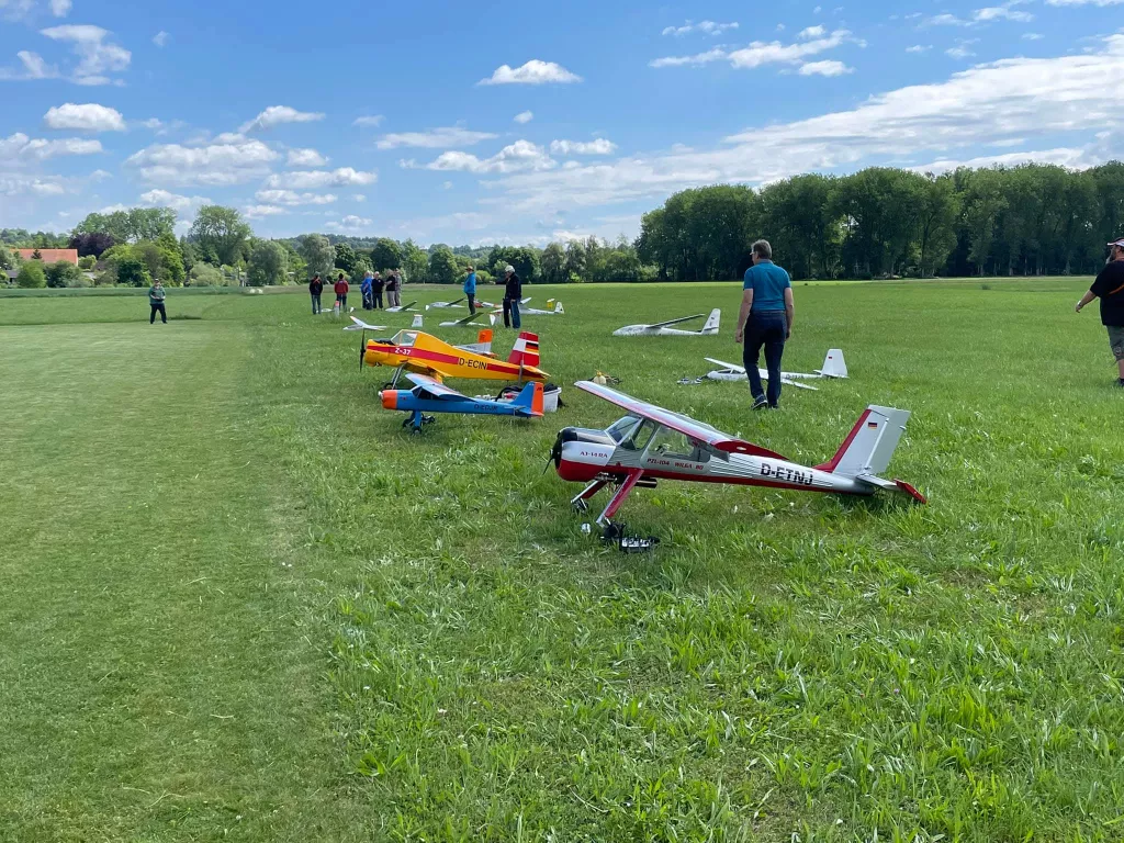 Mehrere Modellflugzeuge in verschiedenen Farben auf einer Wiese, im Hintergrund B&auml;ume und Modellflieger