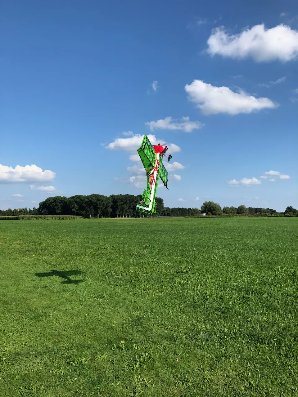 Gr&uuml;n-rotes Modellflugzeug in extremer Schr&auml;glage &uuml;ber gr&uuml;ner Wiese mit blauem Himmel und wei&szlig;en Wolken