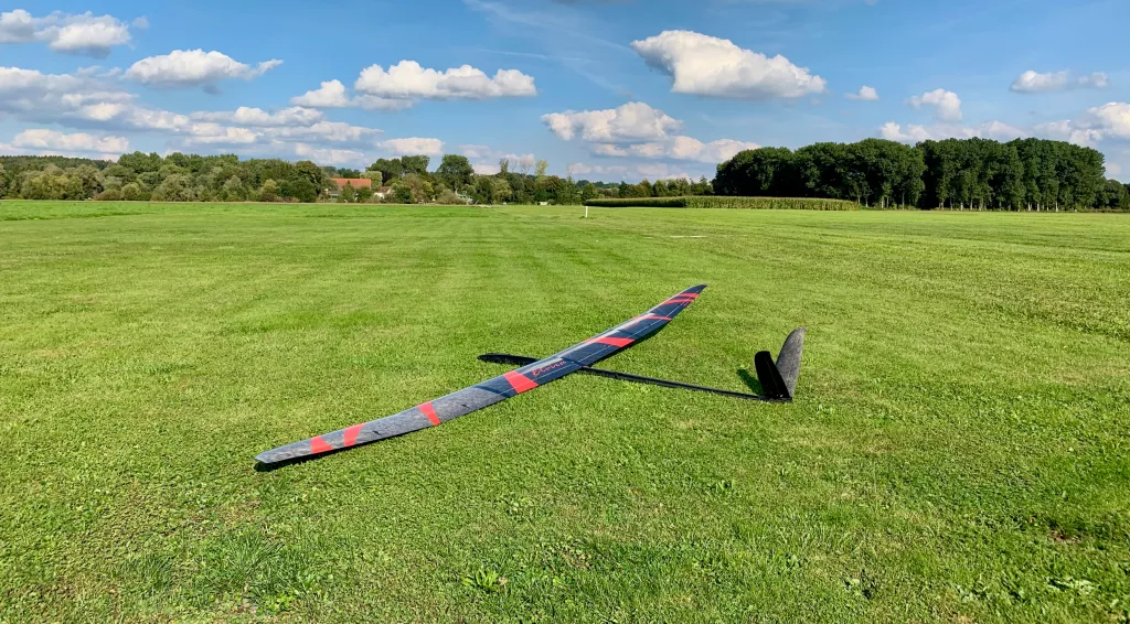 Gro&szlig;es, schlankes Segelflugmodell in grau und rot auf gr&uuml;ner Rasenfl&auml;che, im Hintergrund B&auml;ume und blauer Himmel