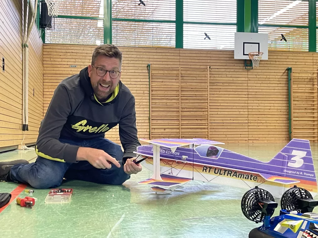 Mann sitzt auf Hallenboden neben einem violetten Modellflugzeug, daneben Zubeh&ouml;r und Werkzeuge