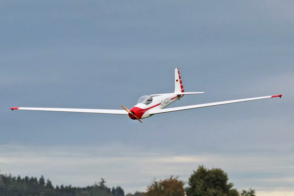 Wei&szlig;es Segelflugmodell mit roten Akzenten fliegt vor grauem Himmel &uuml;ber Baumlandschaft, Kennzeichen D-KOSY sichtbar