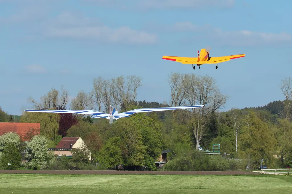 Gelbes Motorflugmodell und blaues Segelflugzeug &uuml;ber gr&uuml;ner Wiese und Baumlandschaft an einem sonnigen Tag